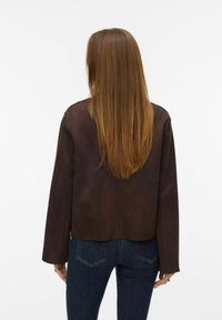 Osoba s dugom ravnom smeškom kestenjaste kose, koja nosi tamnosmeđu jaknu i plave farmerke, stoji okrenuta leđima prema beloj pozadini.