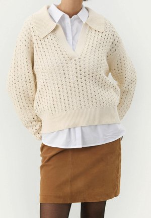 Kvinde iført cremefarvet hæklestrikket sweater over hvid skjorte, parret med brun nederdel og sorte strømpebukser, står med hænderne bag ryggen.