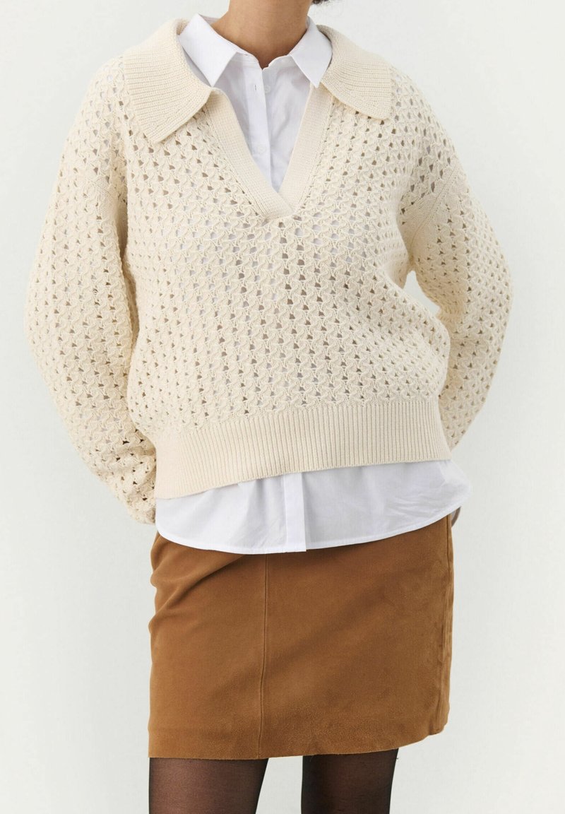 Femme portant un pull en maille crochet crème sur une chemise blanche, associé à une jupe marron et des collants noirs, debout les mains derrière le dos.