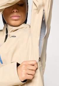 Μπεζ εξωτερικό μπουφάν με μπλε λεπτομέρειες, διαθέτει φερμουάρ στον ώμο και στο πλάι, κατασκευασμένο από μαλακό, υφασμάτινο υλικό, ελαφρύ σχέδιο.