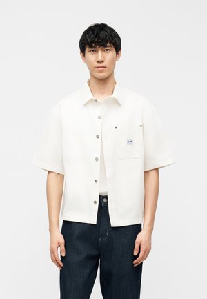 Ung mand med sort hår iført kortærmet hvid skjorte over hvid T-shirt og mørkeblå jeans mod en ensfarvet lys baggrund.