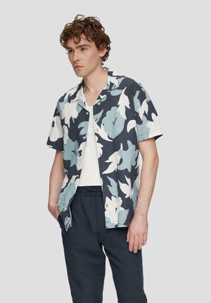 Chemise à manches courtes, à boutons, en tissu bleu marine avec de grands motifs floraux bleu clair et blanc. Portée par-dessus un t-shirt blanc et un pantalon noir.