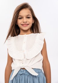 Blusa bianca a balze con punti testurizzati, dotata di un'apertura con bottoni e un punto vita annodato, abbinata a una gonna blu.
