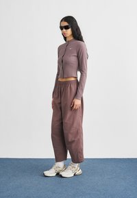 Ριμπ πλεκτή, κοντή μωβ ζακέτα με κουμπιά και λογότυπο· φαρδιά μωβ παντελόνια· λευκά παπούτσια με μπεζ και μαύρες λεπτομέρειες· σκούρα γυαλιά ηλίου.