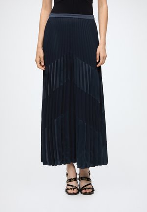 Femme portant une jupe longue plissée taille haute bleu marine foncé, associée à des sandales noires à brides ouvertes avec des clous dorés.