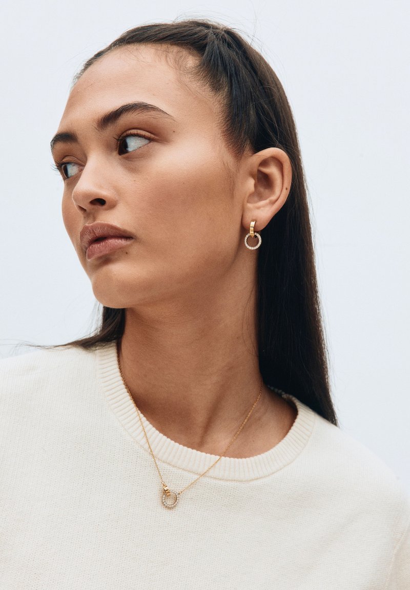Orecchini a cerchio in oro con un anello esterno testurizzato e un cerchio interno liscio. Collana d'oro semplice con un pendente circolare su un maglione chiaro.