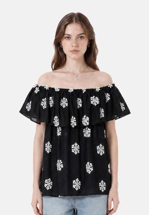 Giovane donna con lunghi capelli castani che indossa un top nero scollato con motivi floreali bianchi e jeans azzurri su uno sfondo semplice.