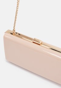 Pochette rectangulaire nude en cuir verni avec cadre en métal doré, fermoir et chaîne délicate sur un fond uni.