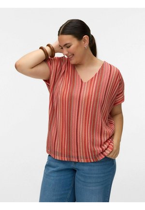 Femme souriante en haut rayé rouge à manches courtes et jean bleu, cheveux attachés en arrière, portant des bracelets en bois et des boucles d'oreilles en argent, posant avec la main sur la tête.