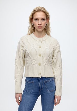 Donna con lunghi capelli biondi che indossa un cardigan color crema lavorato a trecce con bottoni e jeans blu su sfondo bianco neutro.