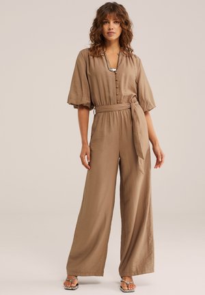 Ženska s kodrastimi lasmi, oblečena v bež široke hlače z ohlapnimi rokavi z napihnjenimi rameni, gumbi spredaj, pasnim trakom in odprtimi sandali.