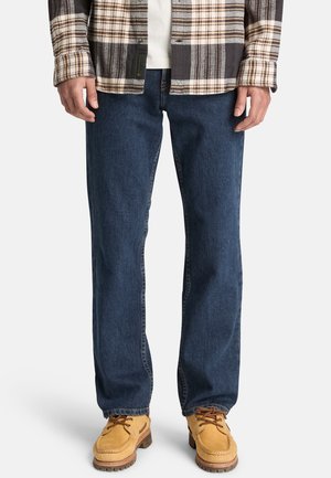 Mann trägt blaue Jeans mit geradem Bein, braune Schnürstiefel, weißes Hemd und braune karierte Jacke, steht mit entspannten Händen.