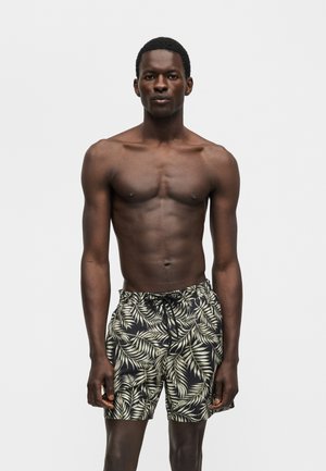 Μυώδης άνδρας στέκεται μπροστά σε μονόχρωμο φόντο, φοράει μαύρο μαγιό με κρεμωτό τροπικό φυλλώδες σχέδιο, τα χέρια χαλαρά δίπλα στο σώμα.