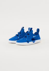 Blaue Slippers mit einer glatten, strukturierten Oberseite, die ausgeschnittene Löcher und weiße Gummisohlen aufweisen. Mit einem Ziehband für einfaches Anziehen.