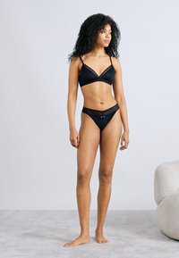 Μαύρο σουτιέν με δαντελένιο τελείωμα και ταίριαστο στρινγκ, με απλό σχεδιασμό και ρυθμιζόμενους ιμάντες, παρουσιάζεται σε μοντέλο πάνω σε ουδέτερο φόντο.