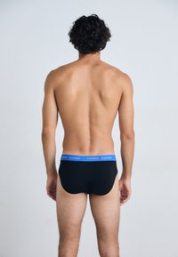 Schwarze Baumwollunterhose mit blauem elastischen Bund, der das Logo "TOMMY HILFIGER" trägt, Ansicht von hinten. Glatte Textur und enganliegende Passform.