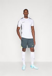 Camisa branca de manga curta com detalhes pretos, calções cinzentos com detalhes em amarelo neón, meias brancas e botas de futebol azul claro. Logotipo da Nike em destaque.