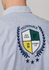 Blauw-wit gestreept shirt met een groot geborduurd patch met groene, gele en witte segmenten, tekst en laurierdetails.