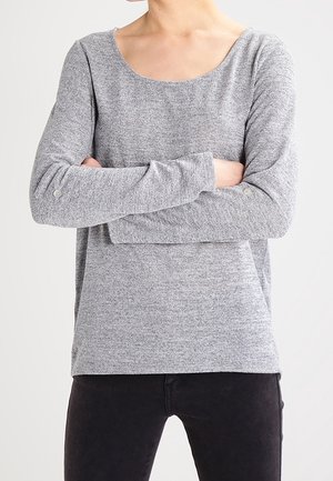 Personne portant un haut en maille gris à manches longues avec des boutons aux poignets, les bras croisés, assorti à un pantalon noir sur un fond uni.