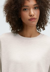 Light beige sweater with a ribbed round neckline. The fabric appears soft with a subtle texture, designed with a relaxed fit.
