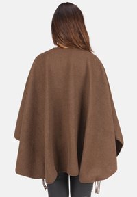 Frau trägt einen lockeren, braunen Wollumhang mit einem schlichten Rücken, der ihren Oberkörper bis zur Hüfte bedeckt, kombiniert mit schwarzen Hosen.