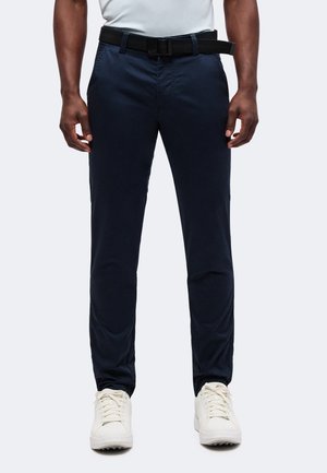Homme portant un pantalon bleu marine foncé avec une ceinture noire et des baskets blanches, debout devant un fond clair uni.