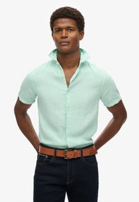 Camicia a maniche corte verde menta chiaro realizzata in tessuto testurizzato, con abbottonatura frontale e colletto. Abbinata a una cintura marrone e jeans scuri.