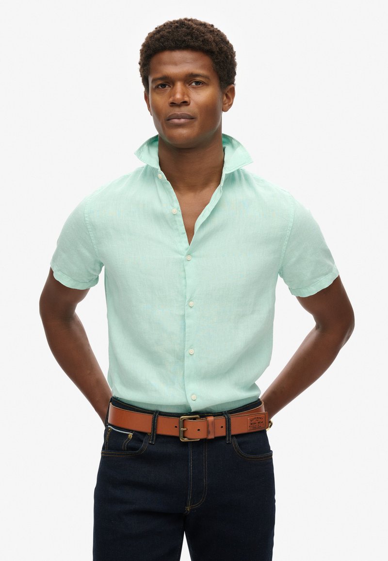 Camicia a maniche corte verde menta chiaro realizzata in tessuto testurizzato, con abbottonatura frontale e colletto. Abbinata a una cintura marrone e jeans scuri.