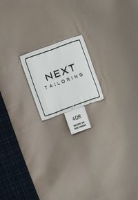 Etiqueta de chaqueta a medida en tela blanca; "NEXT TAILORING" en negro. Talla 40R, con una etiqueta que dice "HECHO EN VIETNAM." Materiales en gris y azul marino.