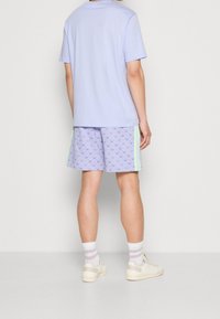 Světle fialové tričko s krátkým rukávem, spárované s vzorovanými fialovými šortkami s zelenými akcenty a jemným designem listů. Bílé ponožky s růžovými pruhy.