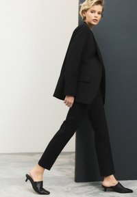 Sort skræddersyet blazer og bukser, glat stof, skarp silhuet. Sammen med spidse sorte mules, blank tekstur. Minimalistisk design.