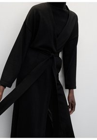 Lang sort kjortel med omslagsdesign, lange ærmer og bindelukning ved taljen. Fremstillet af tekstureret stof med en dyb V-udskæring.