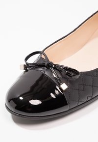 Chaussure plate noire matelassée avec embout en cuir verni brillant et petit nœud décoratif avec embouts métalliques sur fond blanc.