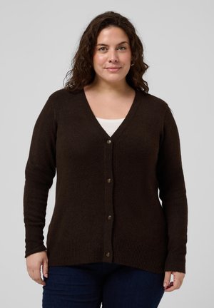 Femme aux cheveux bouclés foncés portant un cardigan marron boutonné par-dessus un haut blanc et un pantalon foncé, debout devant un fond gris.