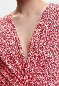 Rode jurk met bloemenpatroon en V-hals, met een geplooid voorpaneel en lichte, gestructureerde stof.