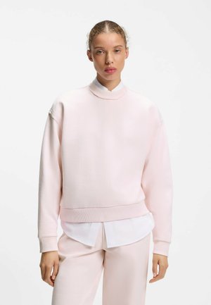 Junge Person mit zurückgebundenem hellen Haar, die einen blassrosa Pullover über einem weißen Hemd und passenden blassrosa Hosen vor weißem Hintergrund trägt.
