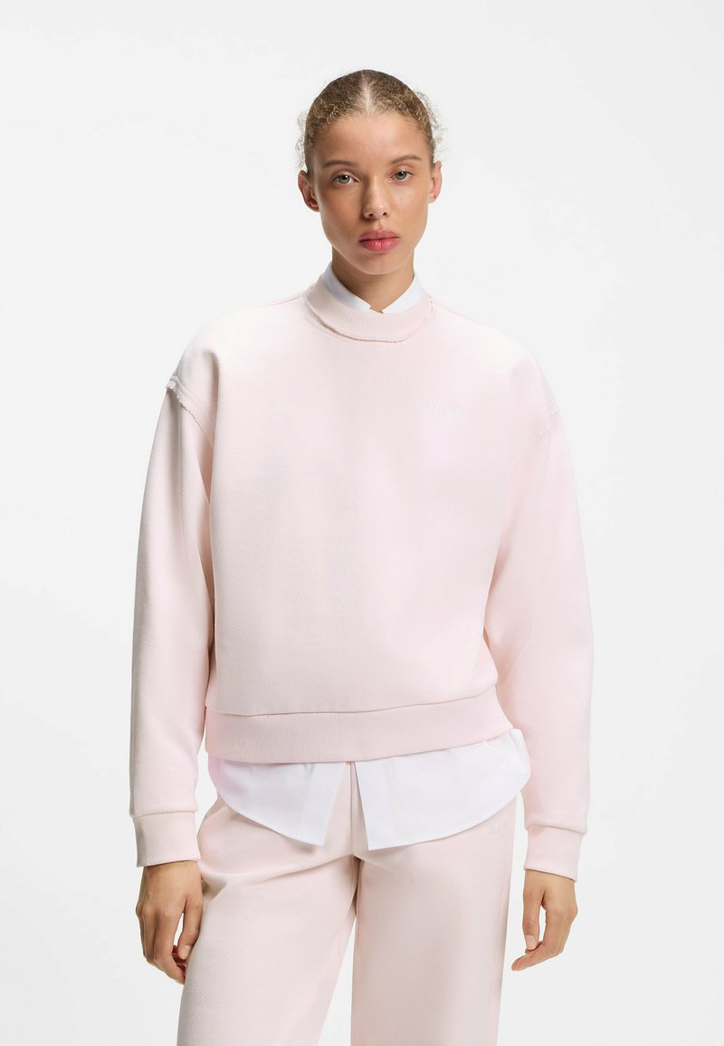 Jeune personne aux cheveux clairs attachés en arrière, portant un sweat-shirt rose pâle par-dessus une chemise blanche et un pantalon rose pâle assorti sur un fond blanc.