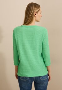 Top verde con texture e maniche a tre quarti, caratterizzato da un taglio rilassato e scollo rotondo, abbinato a jeans blu.