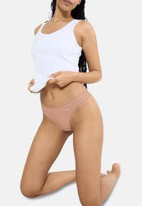 Nude bikini slipjes met kantafwerking en logo, gecombineerd met een witte geribbelde tanktop. Glad materiaal; medium taille pasvorm; minimalistisch ontwerp.