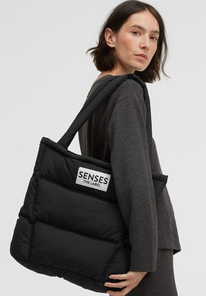 Schwarze gesteppte Tragetasche mit gepolsterter Textur, versehen mit einem weißen Etikett, auf dem "SENSES .THE LABEL" steht, und dicken, schulterfreundlichen Trägern.