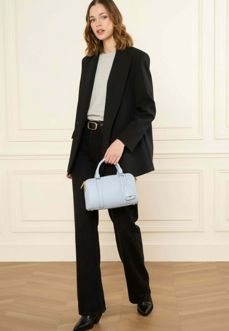 Femme debout à l'intérieur portant un blazer noir, une chemise grise, un pantalon noir, des bottes noires, tenant un sac à main bleu clair avec des accents dorés.