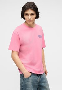 Νεαρός με μεσαίου μήκους σκοτεινά μαλλιά, φοράει ροζ μπλούζα με μπλε γράμματα, χέρια στις τσέπες και ξεθωριασμένο μπλε τζιν.