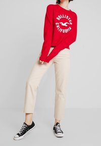 Sudadera roja con "HOLLISTER CALIFORNIA" impreso en blanco, combinada con pantalones beige y zapatillas negras de lona de corte bajo.