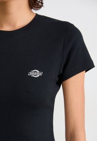 Sort ribbet t-shirt med korte ærmer og rund halsudskæring. Har et lille hvidt logo på venstre brystområde. Glat tekstur.