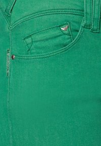 Gröna denimjeans med en framficka som har en distinkt logotyp, subtila sömmar och en mjuk textur. Metalldetalj vid fickan.