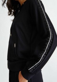 Zwarte zip-up jack met een fitted ontwerp, getextureerde gebreide accenten langs de mouwen en een slanke, minimalistische silhouet.
