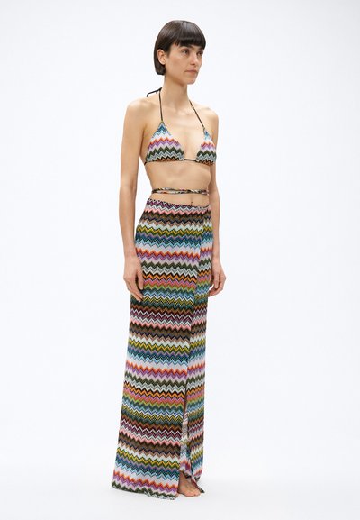 Ženska, oblečena v večbarvni pleteni bikini zgornji del z zig-zag vzorcem ter dolg usklajen ovijalni krilo, stoji bosa pred belim ozadjem.