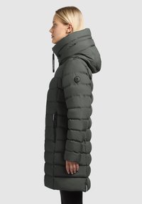 Duga puffer jakna u tamnozelenoj boji, sa uklonjivom kapuljačom, štepanim dizajnom, bočnim džepom sa patentnim zatvaračem i okruglim logotipom na rukavu.
