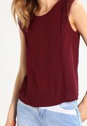 Vrouw met een mouwloze bordeauxrode top met kantafwerking langs de schouders en lichtblauwe spijkerbroek, staand tegen een witte achtergrond.