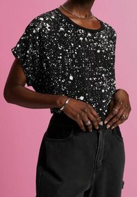 Top negro de manga corta con patrón de salpicaduras blancas, cuello redondo, tejido suave; combinado con pantalones de mezclilla negros de tiro alto. Anillos de plata visibles.